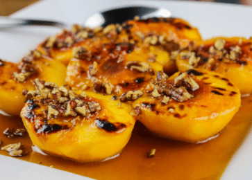 grilled peaches topped with chopped pecans, served with a caramel-like syrup on a white plate. The peaches have a slightly charred, golden appearance from grilling, and the pecans add a crunchy texture on top.