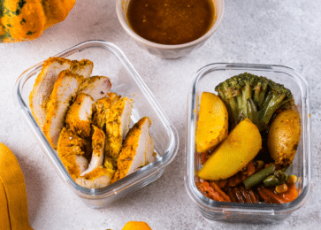 two meal prep containers. One container holds sliced, seasoned turkey breast, while the other contains roasted vegetables, including broccoli, potatoes, carrots, and green beans. A cup of brown sauce is placed beside the containers. The scene is set with a yellow cloth and a small pumpkin in the background.