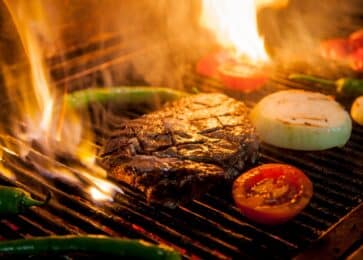 Steak cooking over an open wood fire grill with onions, tomatoes and peppers